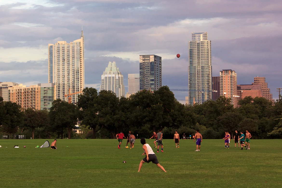 People playing pickup sports on Zilker Park’s open lawn, with a ball in the air and Austin’s skyline behind them